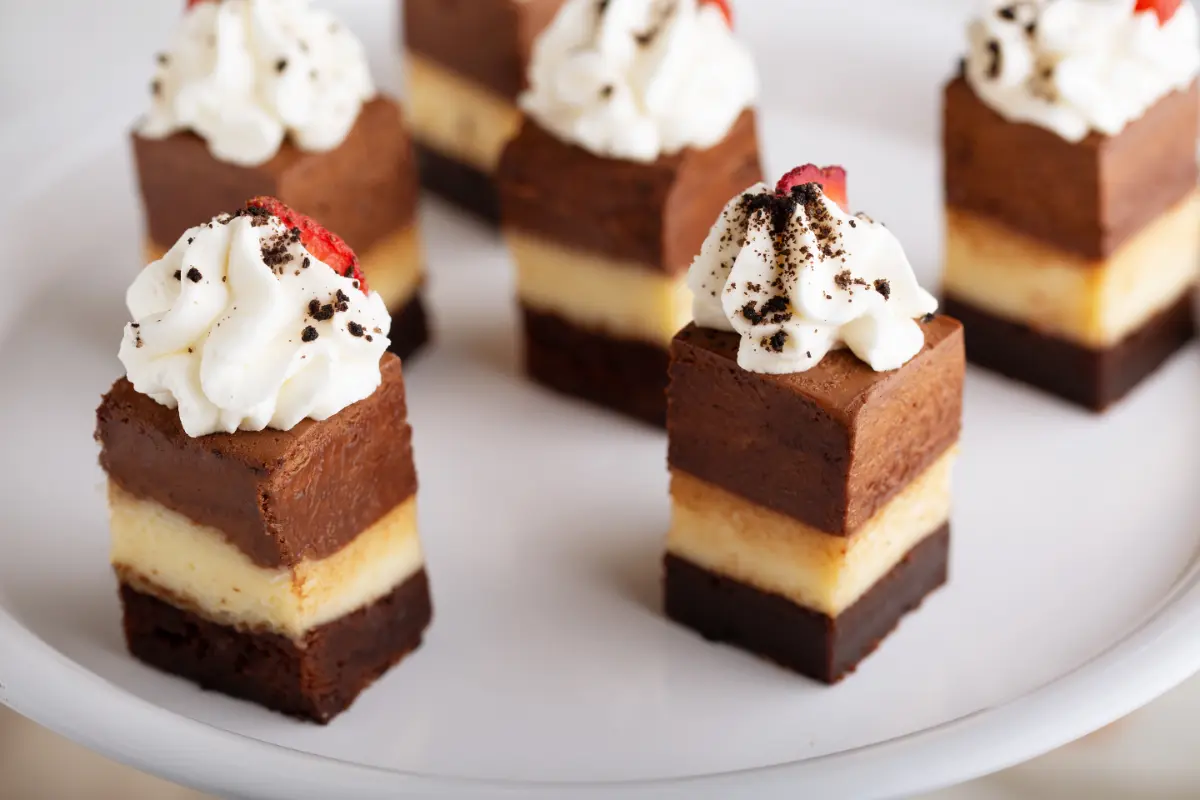 An elegant white plate presents small, layered chocolate mousse cake bites topped with a dollop of whipped cream and a sprinkle of grated chocolate. Each square has a precise layer of cake, mousse, and cream, arranged neatly to showcase the dessert's texture and depth.