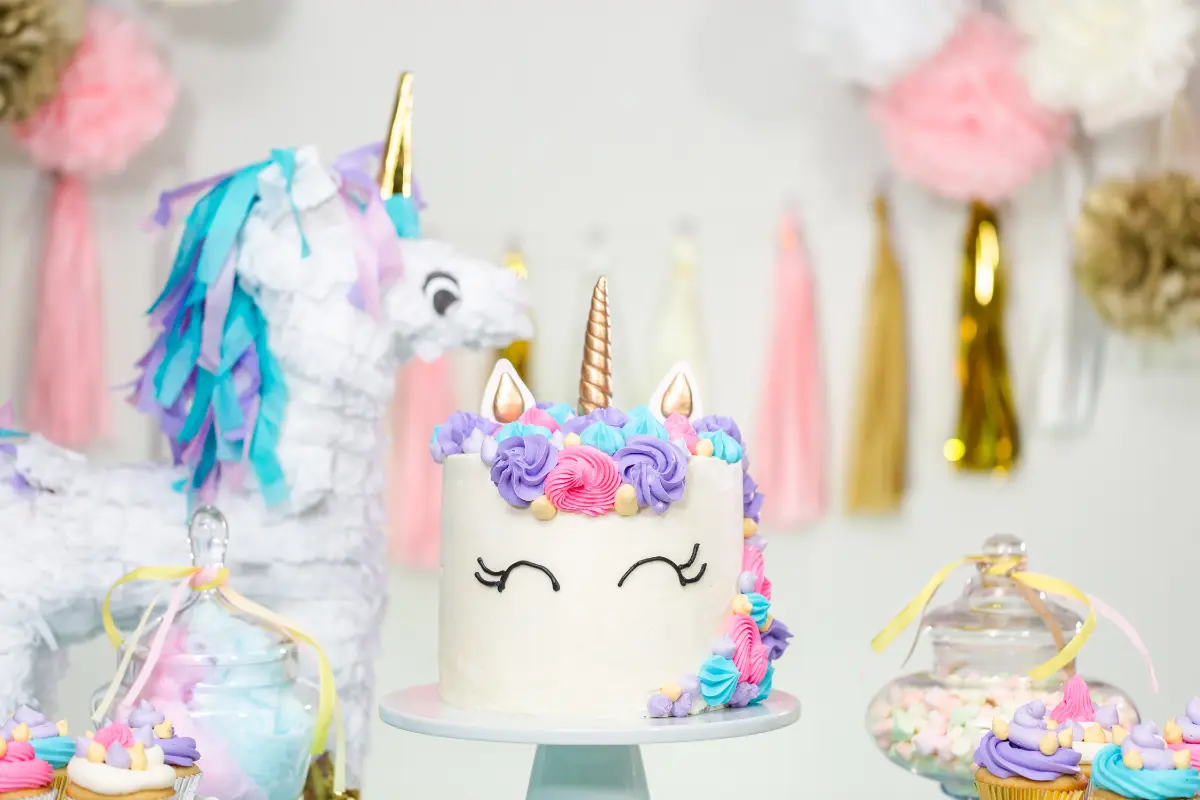 A unicorn cake with pastel frosting and a golden horn, set against a party backdrop with a unicorn piñata and pastel decorations.