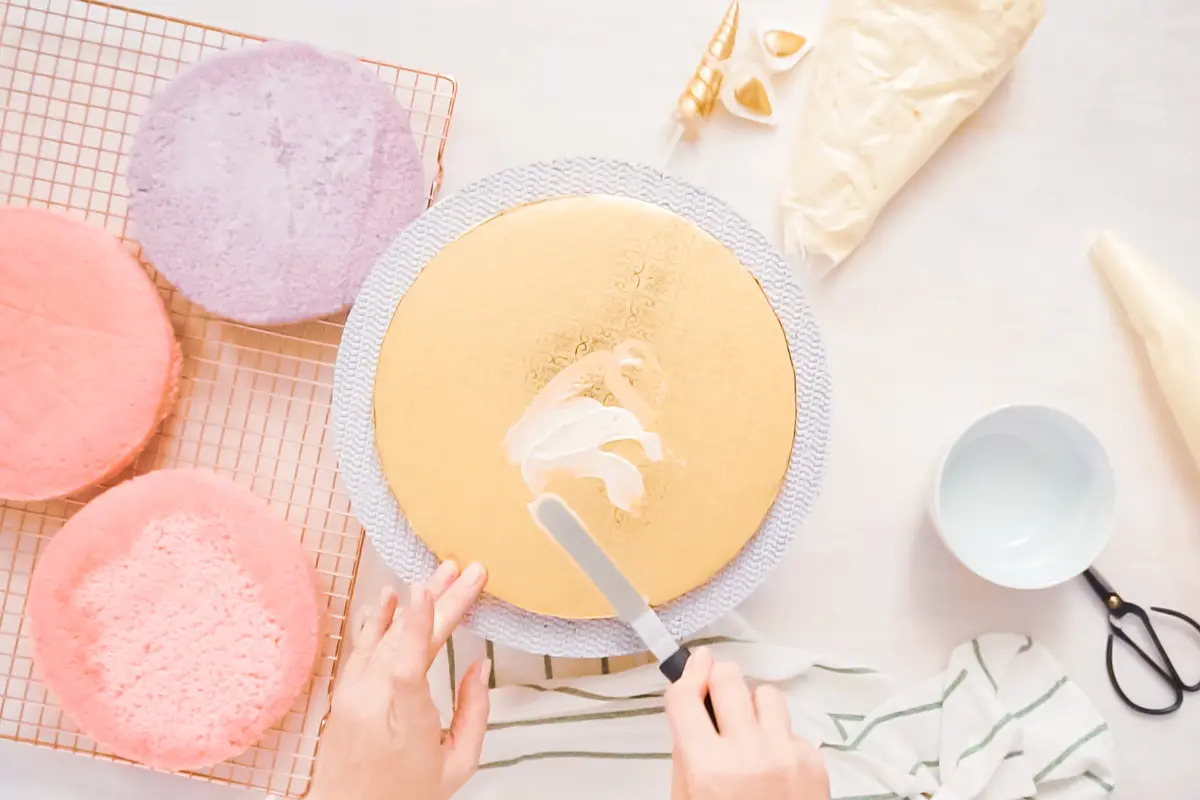 Hands meticulously frosting a vibrant yellow cake layer, with more layers and decorating tools in the background, set for cake assembly.