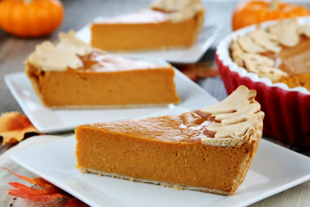 Slices of pumpkin custard pie on plates, with a full pie in the background, embodying the essence of fall's favorite dessert.