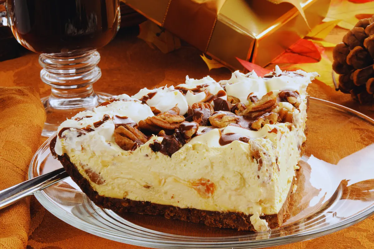 A generous slice of Turtle Cheesecake on a clear plate, garnished with whipped cream, pecans, and chocolate shavings, set against a warm, festive backdrop.