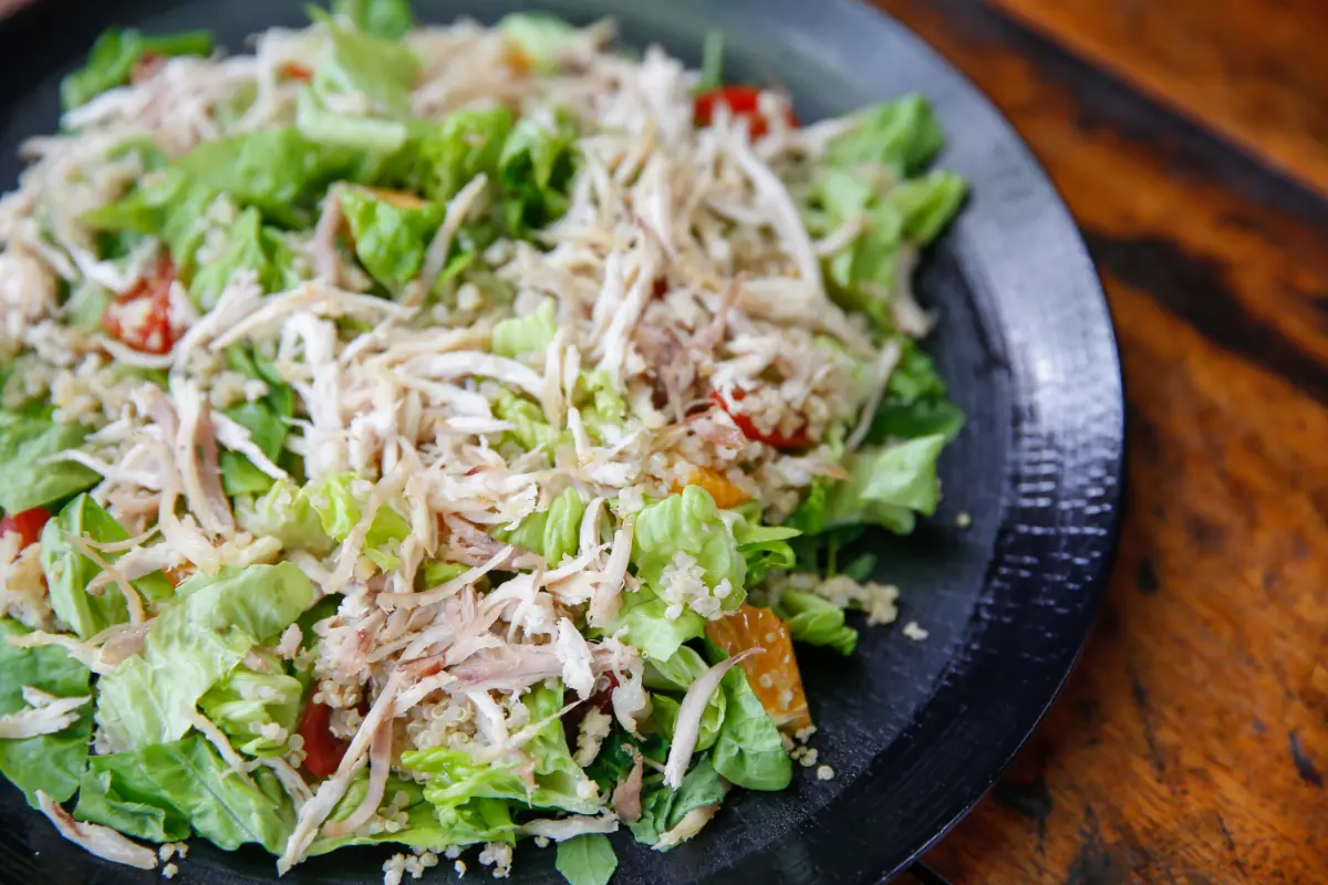 A nutritious salad with shredded chicken, quinoa, lettuce, and tomatoes on a black plate, offering a wholesome meal on a wooden table.