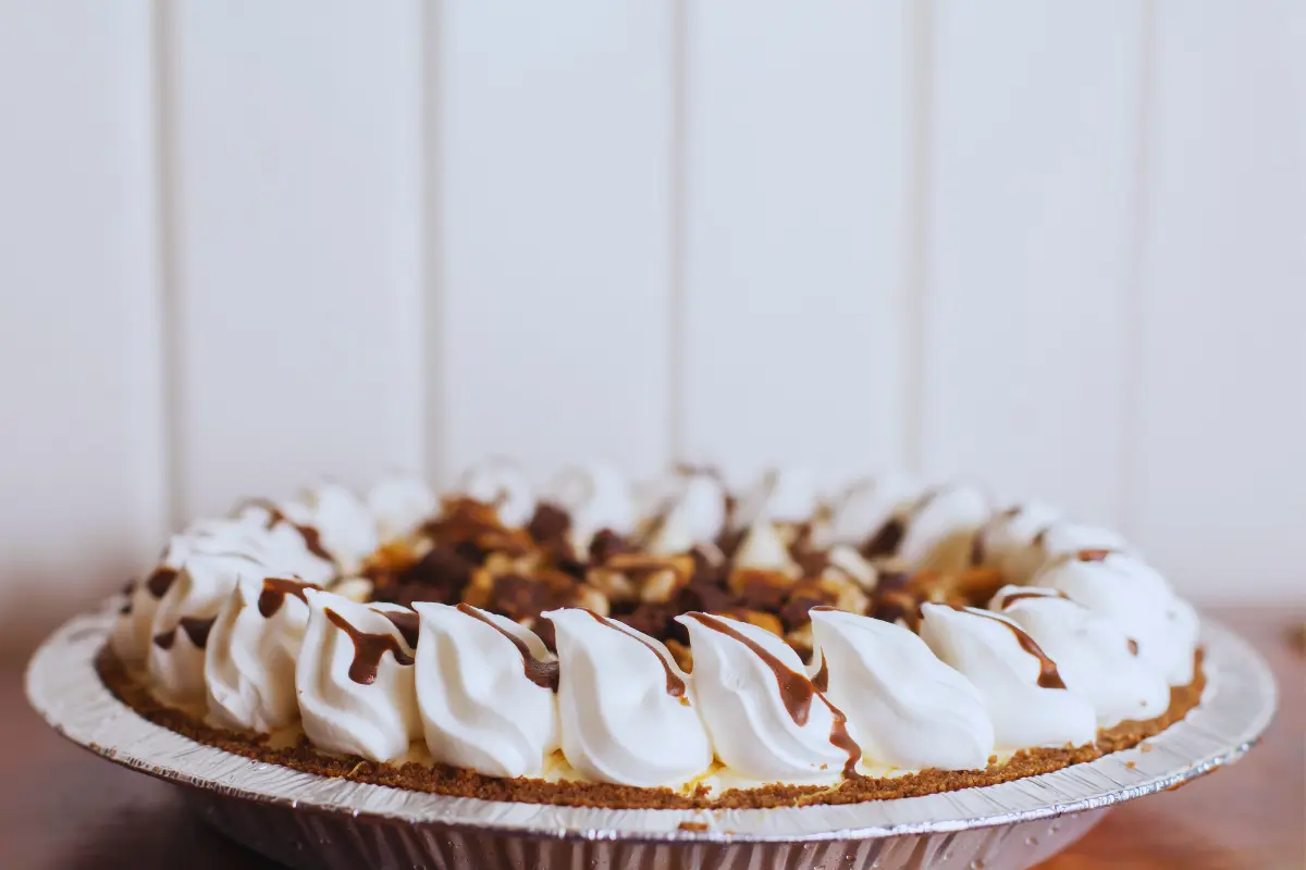 A Turtle Pie in a foil dish, showcasing whipped cream peaks with chocolate drizzle, hinting at the sumptuous caramel and pecan filling within.