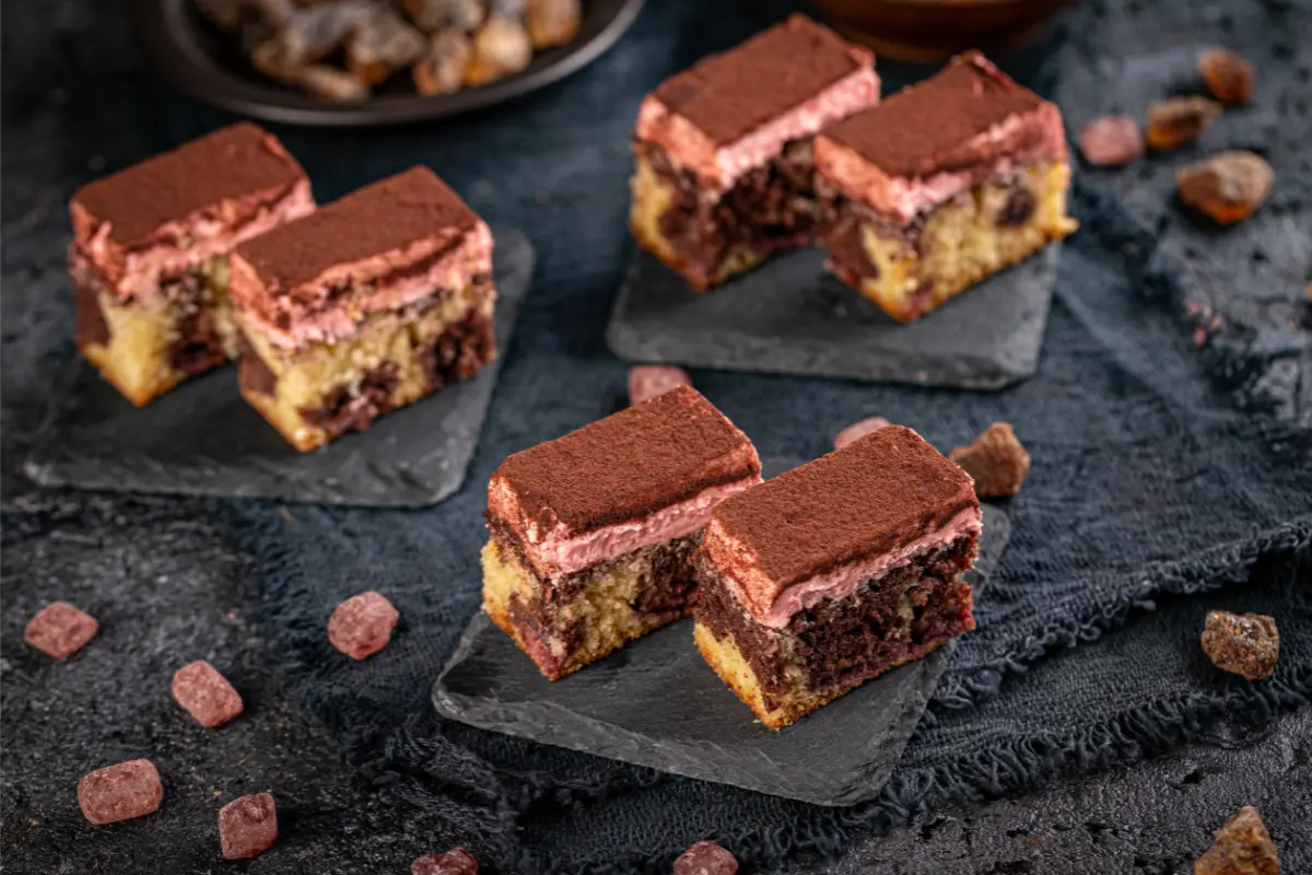 Several small, square tiramisu-inspired cake bites are displayed on a dark slate, dusted with cocoa powder. The contrasting layers of cake and mascarpone cream offer a visual treat, complemented by the rich, dark setting and scattered pieces of chocolate around the slate.