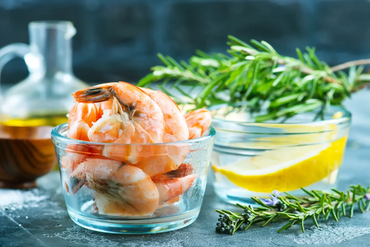 A bowl of cooked shrimp with lemon slices and rosemary in the background, suggesting a flavorful and fresh seafood dish.