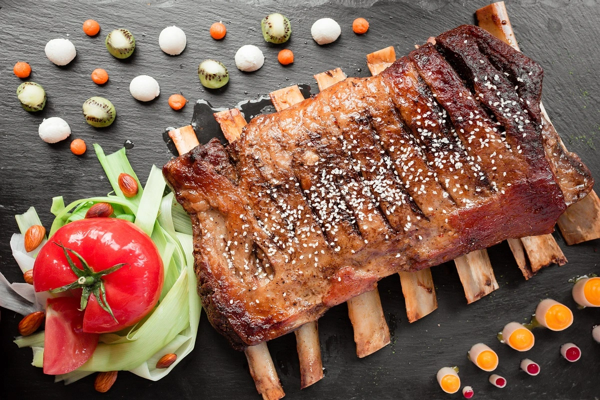 A succulent roast beef ribs adorned with sesame seeds, surrounded by a creative array of colorful vegetables and nuts on a slate background.