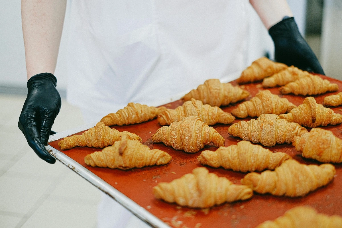 A person in a white apron and black gloves organizing a tray of golden-brown mini croissants.