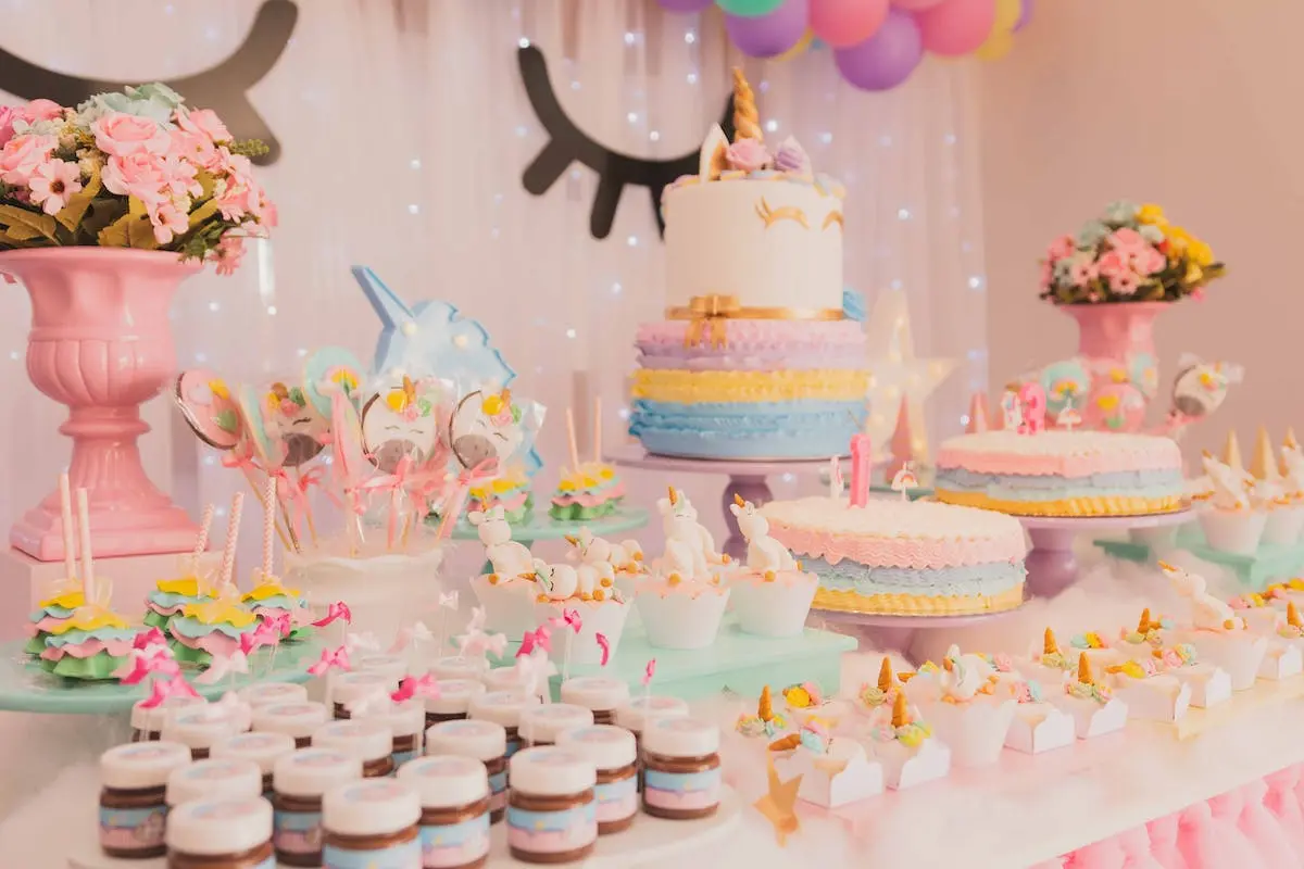 A unicorn-themed party table lavishly decorated with pastel cakes, cupcakes, and floral arrangements, illuminated by twinkling fairy lights.